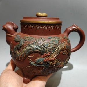 Puede incluir: Tetera de cerámica de color marrón rojizo con un diseño de dragón. La tetera tiene un asa curva, una tapa con un pomo dorado y un pico con forma de cabeza de pájaro. El dragón está detallado con acentos verdes y rojos.