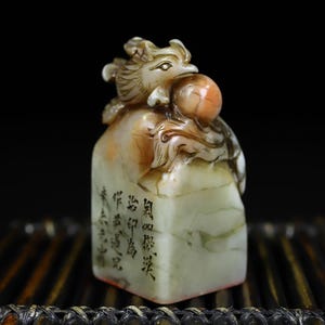May include: A carved stone seal featuring a mythical creature perched atop a rectangular base. The stone is pale green with orange-brown accents. The creature holds a round orange sphere. Black Chinese characters are engraved on the front face.