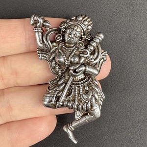 May include: A detailed silver-toned figurine depicting a deity in a dynamic pose, holding a staff and other objects. The figure is adorned with intricate jewelry and a headdress, showcasing fine craftsmanship. The piece has an aged appearance.
