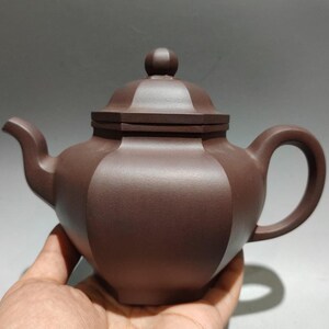 Könnte beinhalten: Eine braune Keramik-Teekanne mit Deckel und Henkel. Die Teekanne hat einen abgerundeten Körper mit geometrischem Design und einen gebogenen Ausguss. Der Griff hat eine Schlaufenform. Der Deckel hat einen kleinen Knopf oben.