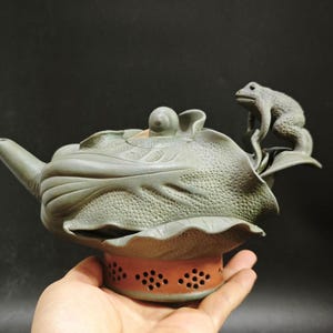 Pode incluir: Uma chaleira de cerâmica com um design de rã e folha. A chaleira é de cor verde suave, com uma rã empoleirada numa folha no topo. A base é de cor castanho-avermelhada com pequenos orifícios.
