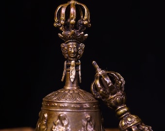 May include: A bronze bell and dorje set. The bell features intricate carvings of figures and patterns, with a detailed crown-like top. The dorje, or thunderbolt, is also bronze and has a similar ornate design. The set is displayed against a dark background.