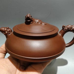 May include: A brown clay teapot with a wide, low-profile body and a flat lid. The lid features a sculpted animal figure as a handle. The spout and the handle are decorated with intricate, carved relief patterns, and the base rests on small feet.
