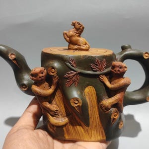 May include: A handcrafted teapot with a tree trunk design, featuring carved monkeys and a small animal on the lid. The teapot has a dark green and brown color scheme, with a curved handle and spout.