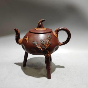 May include: A brown clay teapot with three legs, a curved handle, and a spout. The body features textured, raised branch-like decorations with small circular elements and a small bird figure. The lid has a pointed, curved handle in a matching brown.