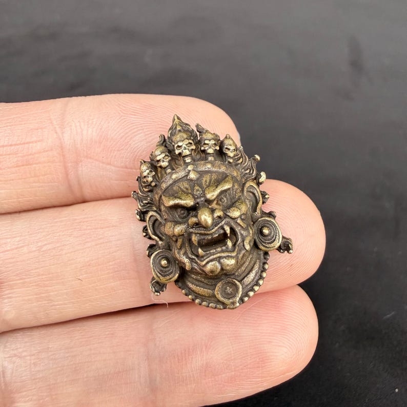 May include: A detailed bronze-colored pendant featuring a fierce, stylized face with multiple skulls atop the head. The pendant has intricate details, including a snarling mouth and circular ear ornaments. The overall design suggests a cultural or historical artifact.