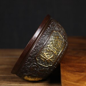 May include: A dark brown, intricately carved, round bowl with gold accents. The bowl has a raised design of stylized flowers and characters in gold.