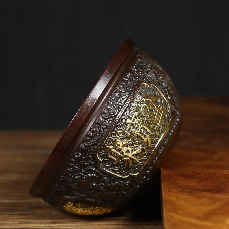 May include: A dark brown, intricately carved, round bowl with gold accents. The bowl has a raised design of stylized flowers and characters in gold.