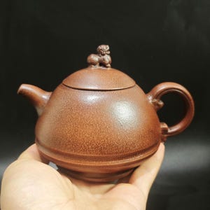 Może przedstawiać: Brązowy ceramiczny imbryk z zaokrąglonym korpusem, zakrzywionym dziobkiem i uchwytem w kształcie pętli. Wieczko zwieńczone jest małą, dekoracyjną figurką. Imbryk trzymany jest w dłoni na ciemnym tle. Imbryk ma około 15 cm długości.