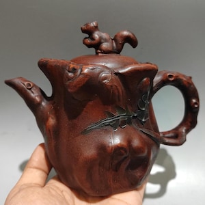 以下が含まれることがあります： 手作りの赤茶色の木製ティーポット。蓋にはリスの置物があります。ティーポットは、曲がった注ぎ口、丸みを帯びたハンドル、彫刻された葉のディテールが特徴です。ティーポットは手で持たれています。