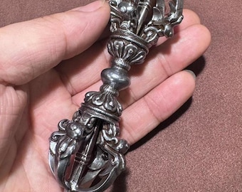 May include: Ornate silver-colored Vajra, a ritual object with intricate details. The Vajra features a central handle with decorative elements and symmetrical, multi-pronged ends. The design includes floral and animal motifs, creating a detailed and symbolic appearance.