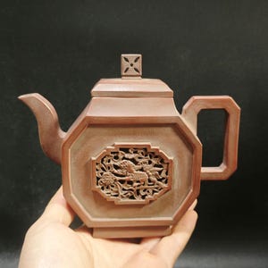 May include: A brown ceramic teapot with an octagonal body and a square handle. The teapot features a decorative carved panel with a mythical creature design. The lid has a small, square finial. The spout is curved.