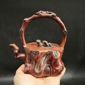Puede incluir: Una tetera artesanal de color marrón rojizo con un diseño orgánico único. La tetera presenta un asa y un pico diseñados para parecerse a ramas de árboles, con una tapa adornada con un detalle esculpido. La tetera mide aproximadamente 15 cm de alto.