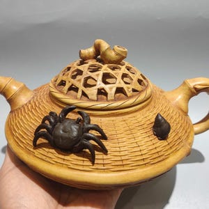 以下が含まれることがあります： 手作りの木製ティーポット。編みかごのデザイン。ティーポットは、格子模様の装飾的な蓋と竹スタイルのハンドルが特徴です。暗いカニが側面に付いています。ティーポットは明るい茶色です。