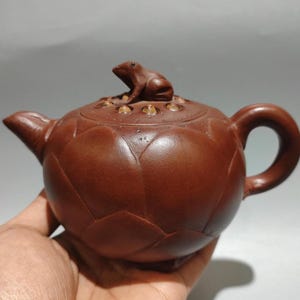 以下が含まれることがあります： 蓋にカエルの置物がある茶色のセラミックティーポット。 ティーポットは、蓮の花のデザイン、湾曲したハンドル、注ぎ口を備えた丸みを帯びたボディをしています。 カエルは蓋の上に止まっています。