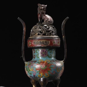 May include: An antique bronze incense burner with a detailed floral design on a turquoise base. The burner features a red band with Chinese characters and a decorative lid topped with a small bronze lion. Two curved handles extend upwards.