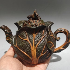 Può includere: Una teiera artigianale dettagliata con un design a forma di foglia. Il corpo verde scuro presenta prominenti venature arancioni-marroni e una superficie strutturata. Il manico e il beccuccio sono curvi e il coperchio è sormontato da un elemento decorativo.
