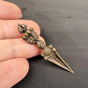 May include: A bronze-colored, ornate dagger with a detailed handle featuring carved faces and a crown-like top. The blade is triangular and tapers to a point. The dagger is held in a hand against a dark background.