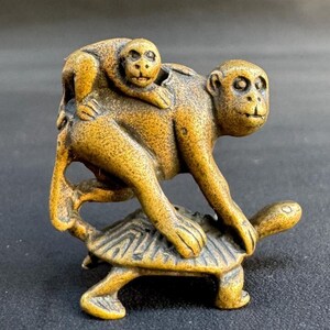 Op de afbeelding: Een gedetailleerde sculptuur van een aap die een kleinere aap op zijn rug draagt, bovenop een schildpad. De sculptuur heeft een warme, goudbruine kleur, met ingewikkelde details op de vacht van de aap en het schild van de schildpad.