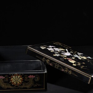 May include: A rectangular black lacquer box with a removable lid. The lid features an inlaid mother-of-pearl design of a bird perched on a vine with leaves. The front of the box base displays a colorful floral motif with gold-toned accents.