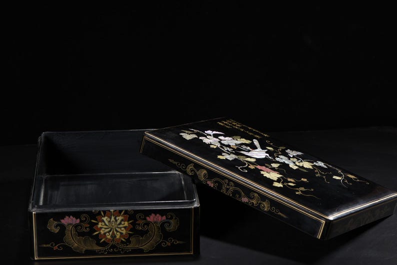 May include: A rectangular black lacquer box with a removable lid. The lid features an inlaid mother-of-pearl design of a bird perched on a vine with leaves. The front of the box base displays a colorful floral motif with gold-toned accents.