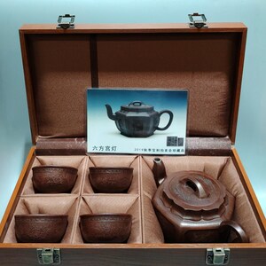 Puede incluir: Un juego de té de madera marrón oscuro con una tetera y cuatro tazas, presentado en una caja de madera con interior marrón. También se incluye una pequeña imagen de la tetera. La caja tiene cierres metálicos.