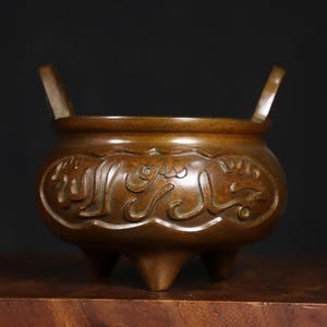 May include: Bronze-colored incense burner with three legs and two handles. The vessel features ornate, stylized Arabic script around the body. The burner sits on a wooden surface against a dark background.