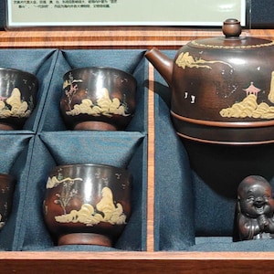 May include: A wooden tea set with a teapot, four cups, and a small statue of a laughing Buddha. The teapot and cups are brown with gold landscape designs. The set is displayed in a wooden box with a blue interior.