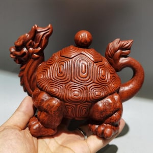 Puede incluir: Escultura de madera marrón rojiza detallada de una criatura parecida a una tortuga con rasgos de dragón. La escultura tiene un caparazón texturizado, una cabeza de dragón y una cola curvada. La tapa tiene un pequeño pomo redondo. La escultura se sostiene en una mano.