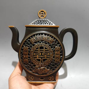 Puede incluir: Una tetera marrón oscuro con intrincados grabados dorados y un diseño circular en la parte delantera. La tetera presenta un asa, un pico y una tapa decorativa. El diseño incluye caracteres y símbolos chinos.