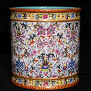 以下が含まれることがあります： 白を基調とした円筒形の陶器の花瓶で、ピンク、黄色、青、緑の鮮やかな花柄で飾られています。 上部と下部の縁には、複雑なデザインの装飾的なバンドがあります。 内側は水色です。
