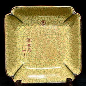 Może przedstawiać: Kwadratowy, żółty ceramiczny talerz z wzorem spękanego szkliwa. Talerz ma złoty brzeg i czarne akcenty na rogach. Zawiera złote detale i czerwone japońskie znaki.