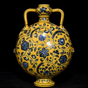 Pode incluir: Um vaso de cerâmica amarelo com padrões florais azuis. O vaso tem duas alças e um corpo redondo. O design floral cobre toda a superfície do vaso, com um padrão detalhado de flores e folhas. O vaso assenta numa pequena base decorativa.