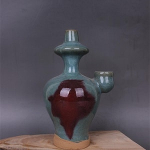 May include: A green and brown ceramic vase with a spout and a unique design. The vase has a glaze that creates a mottled effect.