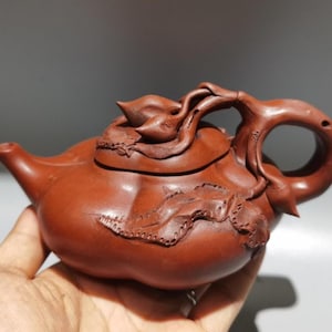 Puede incluir: Una tetera de cerámica marrón rojizo con intrincados diseños florales y de hojas. La tetera tiene un asa curva y un pico, con una tapa. La tetera se sostiene en una mano.