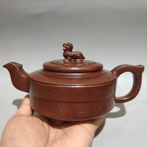 May include: A dark brown clay teapot with a flat, cylindrical body and a curved handle. The lid features a small, sculpted mythical creature as a handle. The exterior of the teapot is decorated with engraved characters around the middle section.