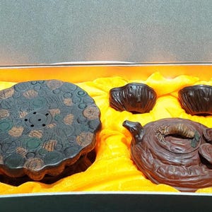 May include: A dark brown tea set consisting of a round tray with a textured circular pattern, two small cups, and a teapot with a sculpted handle and lid. The items are arranged inside a rectangular box lined with bright yellow fabric.
