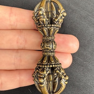 Könnte beinhalten: Verziertes Vajra in Bronzefarbe, ein Ritualobjekt mit komplizierten Details. Das doppelseitige Zepter weist dekorative Elemente auf, darunter drachenartige Figuren und einen zentralen Griff. Das Design ist symmetrisch und detailliert, mit einem spitzen Ende.