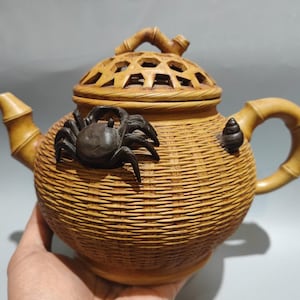 以下が含まれることがあります： 織りかごのデザインの装飾的なティーポット。 ティーポットはタン色で、竹のハンドルと注ぎ口が付いています。 濃い茶色のカニとカタツムリが側面に付いています。 蓋はハニカム模様です。