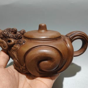 Könnte beinhalten: Braune Keramik-Teekanne mit einem Drachenkopf-Ausguss und einem gebogenen Griff. Die Teekanne hat einen Deckel mit einem kleinen Knopf und ein Wirbeldesign auf dem Körper. Die Teekanne wird in einer Hand gehalten.