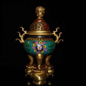 Puede incluir: Un quemador de incienso ornamentado con acabado dorado. El quemador tiene un cuerpo turquesa con diseños florales en rosa, amarillo y blanco. La tapa es roja y dorada, con intrincados patrones calados. Tiene tres patas y dos asas.