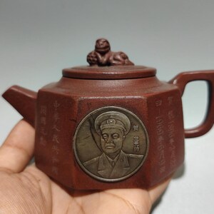 Könnte beinhalten: Eine sechseckige Teekanne aus braunem Ton mit einem runden Metallmedaillon auf der Vorderseite, das eine uniformierte Person zeigt. Der Deckel ist mit einer kleinen, geformten Tierfigur verziert. Chinesische Schriftzeichen sind in die Seiten eingraviert.