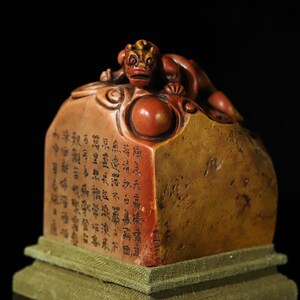 May include: A rectangular stone seal featuring a carved mythical creature on top and a smooth sphere. The side displays vertical columns of black Chinese calligraphy characters. The stone has a warm, mottled tan and reddish-brown colour palette.