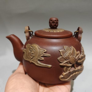 Puede incluir: Una tetera de arcilla marrón con un pico curvo, una tapa con un pomo texturizado y dos enganches metálicos para el asa. El cuerpo está decorado con relieves de una flor de loto y un emblema circular con caracteres chinos en color dorado.