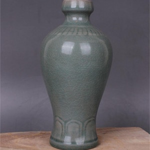 May include: A celadon-colored ceramic vase with a crackle glaze finish. The vase has a rounded body, a long neck, and a flared rim. The base features a decorative pattern. The vase is displayed on a wooden surface.