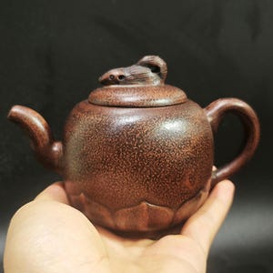 以下が含まれることがあります： テクスチャ加工された表面と小さなハンドルが付いた、茶色のセラミックティーポット。蓋には装飾的な要素があります。ティーポットは暗い背景に対して手で持たれています。ティーポットは、お茶を淹れるように設計されています。