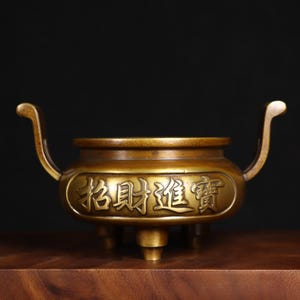 May include: A bronze incense burner with curved handles and three small feet. The vessel features embossed Chinese characters. The burner has a rounded body and a warm, metallic finish, suggesting a traditional or decorative purpose.