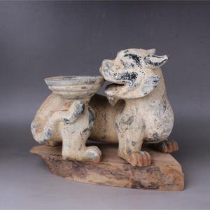 May include: A white ceramic sculpture of a mythical lion with a black and white painted design. The lion is holding a small, white ceramic bowl. The sculpture is sitting on a wooden base.