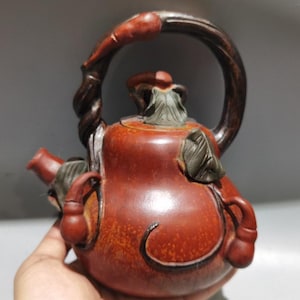 Może przedstawiać: Czerwono-brązowy czajnik z zakrzywionym uchwytem i wylewką. Czajnik ma kształt tykwy z dekoracyjnymi akcentami liści i winorośli. Uchwyt jest ciemnobrązowy, a akcenty jaśniejsze.