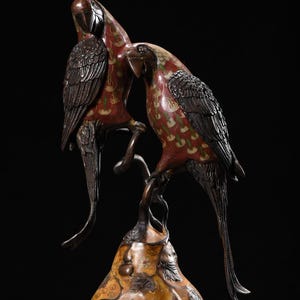 Peut inclure: Une sculpture en bronze de deux perroquets perchés sur une branche, au sommet d'une base en forme de poire. Les perroquets ont un plumage à motifs rouges et dorés, avec des ailes et des becs sombres. La base est décorée de motifs floraux.